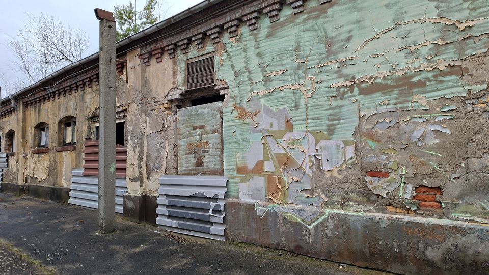 Verwitterte Außenwand eines alten Industrie- oder Lagergebäudes mit bröckelndem Putz, freiliegendem Mauerwerk und verblassten Graffiti. Entlang der Fassade stehen ein Betonmast und gestapelte Dämmplatten; kleine Fensteröffnungen und eine Lüftungsöffnung sind zu sehen.