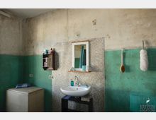 Abgenutztes Badezimmer mit türkisgrünen Wänden und deutlichen Gebrauchsspuren. Im Zentrum ein Waschbecken mit Spiegel, daneben eine kleine Wandablage, ein Staubwedel und eine Bürste an der Wand aufgehängt.