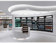Innenraum einer modernen Apotheke mit geschwungener Decke und beleuchteten, schwarzen Regalen, die eine Vielzahl von Produkten in verschiedenen Farben präsentieren. Im Vordergrund befinden sich runde, weiße Verkaufstische mit integrierten Displays.