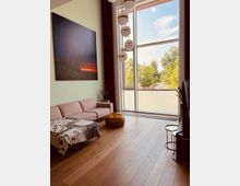 Wohnzimmer mit hohen Decken, einem großen Fenster, das auf einen grünen Außenbereich blickt, und moderner Einrichtung. Im Raum befinden sich ein rosafarbenes Sofa, ein kleiner runder Holzhocker, ein Teppich mit dekorativem Muster und eine hängende Lampe mit mehreren runden Elementen.