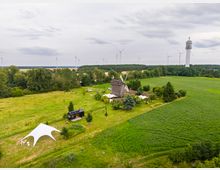 Luftaufnahme einer ländlichen Landschaft mit Feldern und Baumgruppen, in deren Mitte eine historische Windmühle mit Nebengebäuden und weißen Zeltdächern steht. Am Horizont sind mehrere Windkraftanlagen sowie ein hoher Aussichtsturm am Waldrand zu sehen.