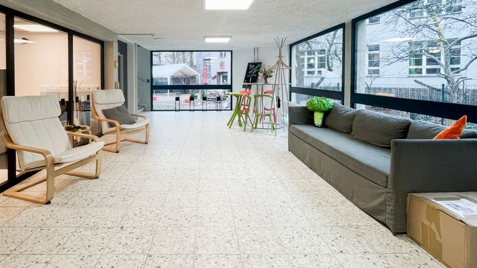 Heller Aufenthaltsbereich in einem Gebäude mit großflächigen Fensterfronten, gefliestem Boden und Deckenleuchten. Links stehen zwei helle Sessel vor Glaswänden, rechts ein graues Sofa; im Hintergrund ein kleiner Stehtisch mit Hockern und Blick in einen Hofbereich.