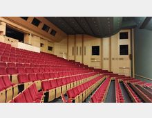 Ein moderner Hörsaal mit ansteigenden Reihen aus roten gepolsterten Sesseln und Holzwänden. Die Decke ist grün mit eingebauten Beleuchtungselementen, und an den Seiten befinden sich Türen und Fenster.