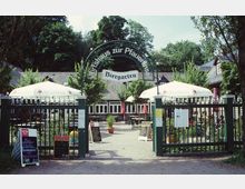 Eingezäunter Biergarten mit dem Schriftzug „Wirtshaus zur Pfaueninsel“ über dem Eingang. Im Hintergrund stehen Tische und Stühle unter großen weißen Sonnenschirmen, umgeben von Bäumen und Gebäuden mit dunklen Dächern.