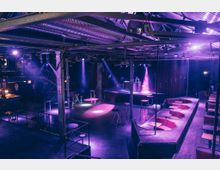 Moderner Veranstaltungsraum mit Bühne und DJ-Pult, beleuchtet durch lila und pinke Scheinwerfer, umgeben von Sitzbereichen mit roten Polstern sowie Stehtischen. Die Decke zeigt freiliegende Rohre und Stahlkonstruktionen, die den industriellen Charakter des Raumes betonen.