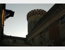 Blick auf einen historischen Burgturm aus Ziegelstein mit zinnenförmigem Abschluss, umgeben von massiven Burgmauern mit kleinen Fenstern. Der Himmel im Hintergrund ist hell, während der Vordergrund im Schatten liegt.