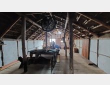 Holzinterior eines historischen Dachbodens mit sichtbarem Gebälk und schrägem Dach. In der Mitte steht ein rustikaler Tisch, umgeben von einfachen Möbeln und Gegenständen, die eine altertümliche Atmosphäre vermitteln.