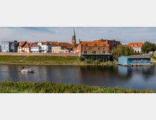 Stadtansicht mit einer Reihe von Häusern in bunten Farben und roten Dächern, die an einem Fluss mit begrüntem Ufer liegen; im Hintergrund ein Kirchturm mit grüner Spitze und rechts ein blaues Bootshaus. Ein kleines Motorboot fährt auf dem Fluss.