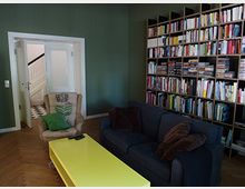 Wohnzimmer mit dunkelgrüner Wand, einem großen Bücherregal, einem blauen Sofa, einem beigefarbenen Sessel und einem gelben Couchtisch. Im Hintergrund ist eine Tür, die zu einem Flur mit schwarz-weiß kariertem Boden führt.