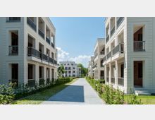 Moderne Wohnanlage mit hellen, mehrstöckigen Gebäuden und Balkonen auf beiden Seiten eines gepflasterten Weges. Zwischen den Gebäuden befinden sich gepflegte Grünflächen mit kleinen Hecken und einzelnen Bäumen, im Hintergrund weitere Wohngebäude und ein klarer Himmel.