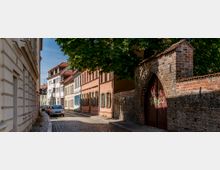 Schmale gepflasterte Gasse mit historischen Wohnhäusern und farbigen Fassaden auf der linken Seite, daneben eine alte Ziegelmauer mit einem großen, hölzernen Tor in gotischem Bogenstil. Im Hintergrund stehen parkende Autos, und Bäume spenden Schatten.