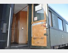 Außentür eines historischen Zugwaggons mit Holzverkleidung und Hinweisschild „Nicht öffnen, bevor der Zug hält“. Der Blick führt ins Innere des Waggons mit weiteren Holzpaneelen.