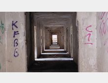 Verlassener, stark heruntergekommener Gebäudekorridor mit mehreren hintereinanderliegenden offenen Türrahmen. Die Wände sind mit Graffiti bedeckt, der Boden ist schmutzig, und am Ende des Korridors fällt Licht durch eine Öffnung.