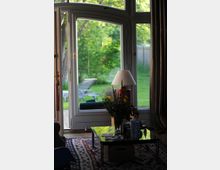 Wohnzimmeransicht mit einer großen Glastür, die zu einem grünen Garten mit Rasen und Liegestühlen führt. Im Vordergrund steht ein niedriger Glastisch mit Blumenstrauß, darauf liegt ein gemusterter Teppich.