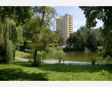 Hochhaus im Hintergrund eines grünen Parks mit einem kleinen See im Vordergrund, umgeben von Bäumen und Grasflächen. Die Umgebung wirkt dicht bewachsen, mit hängenden Ästen und einem geschwungenen Uferweg entlang des Wassers.
