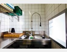 Kleine Industrieküche mit weiß gefliesten Wänden, einem Edelstahlspülbecken mit flexiblem Wasserhahn, einem Schneidebrett auf der Arbeitsfläche und Regalen mit Kisten an der Wand. Rechts befindet sich ein großes Fenster, das Licht hereinlässt.