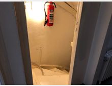 Blick auf eine schmale, weiße Wendeltreppe in einem engen, beleuchteten Bereich mit weiß gestrichenen Wänden; ein roter Feuerlöscher hängt an der Wand, und lose Kabel liegen am Boden.