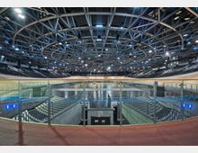 Innenansicht einer großen Radrennbahn-Halle mit ovaler Holzbahn, dunklen Tribünenreihen und einer zentralen Infield-Fläche. Über dem Innenraum spannt sich eine komplexe Stahlträger-Dachkonstruktion mit ringförmigen Beleuchtungselementen und vielen Scheinwerfern.