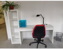 Ein heller Arbeitsbereich mit weißem Schreibtisch, schwarzem Drehstuhl mit roter Umrandung und einer schwarzen Schreibtischlampe. Daneben befindet sich eine weiße Regalwand mit einer Topfpflanze oben und blauen Ablagefächern auf dem Schreibtisch; im Hintergrund ist ein Flur erkennbar.