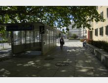 U-Bahn-Eingang "Platz der Luftbrücke" mit gläsernem Überdachungsbau, umgeben von einem Gehweg mit Bäumen im Vordergrund. Im Hintergrund sind moderne Gebäude und eine ruhige Straße erkennbar, eine Person fährt Fahrrad und eine andere sitzt auf einer Steinbank.