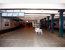 Eine U-Bahn-Station mit sichtbaren Stahlträgern und weißen Wänden, die mit Werbeplakaten dekoriert sind. Im Vordergrund stehen Sitzbänke, während eine Uhr und ein beleuchtetes Schild von der Decke hängen; der Bahnsteig ist gut beleuchtet und leer.