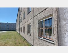 Ein längliches, mehrstöckiges Plattenbaugebäude mit grober Putzfassade und Fenstern mit rostigen Gitterstäben; im Hintergrund ein weiteres ähnliches Wohngebäude und eine grasbewachsene Freifläche unter klarem, blauem Himmel.