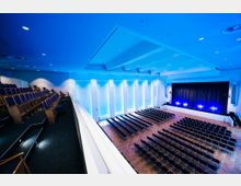 Moderner Veranstaltungssaal mit einer Bühne, davor Sitzreihen auf einem Parkettboden und einem Balkonbereich mit gepolsterten Sitzen. Wände und Decke sind in Blau beleuchtet, große Wandpanele betonen die Höhe des Raumes.