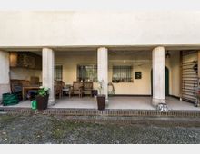 Überdachter Außenbereich eines Gebäudes mit drei großen weißen Säulen, gepflastertem Boden und dekorativen Blumentöpfen. Im Hintergrund sind eine Sitzgruppe aus Holz, ein Wandregal, Gitterfenster und diverse Gartengeräte zu sehen.