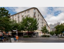 Ecke einer Wohnstraße mit einem mehrstöckigen Wohnhaus im Altbaustil, großen Bäumen und kleinen Geschäften im Erdgeschoss. Im Vordergrund sind Straßenschilder, ein Hund auf einer Bank und geparkte Autos zu sehen.