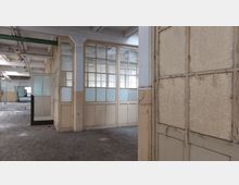 Ein leerer, verfallener Raum in einem industriellen Gebäude mit Glastrennwänden, teilweise matten Fensterscheiben und abblätternder Farbe. Der Holzboden ist staubig, und Rohre sowie Leitungen sind an der Decke sichtbar.