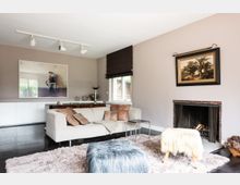 Wohnzimmer mit beige gestrichenen Wänden, einer hellen Couch mit Lederkissen, einem flauschigen Teppich und Hockern mit Textilbezügen. Im Raum befinden sich ein Kamin mit Backsteinrahmen, ein großer Fotodruck, ein Landschaftsgemälde und ein Fenster mit braunem Rollo, das Tageslicht hereinlässt.