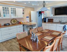 Wohnküche mit hellen, klassischen Küchenmöbeln, einer Arbeitsplatte aus Holzoptik und eingebautem Waschbecken. Im Vordergrund steht ein Holz-Esstisch mit Platzdeckchen und gepolsterten Stühlen; an der Decke eine Pendelleuchte.