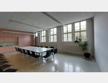 Heller Besprechungsraum mit rechteckigem Tisch und schwarzen Stühlen, großen Fenstern an der rechten Wand, einer Küche mit rosa Schränken links und einer Zimmerpflanze in einer Ecke.