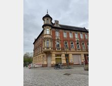 Eckgebäude im historistischen Stil mit roten Ziegelwänden und gelb-beigen Fassadenelementen, darunter ein runder Erker mit Turmhaube. Im Vordergrund eine Kopfsteinpflasterstraße, ein paar Fahrräder und einige junge Bäume.