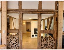 Innenraum eines rustikalen Gebäudes mit freiliegenden Holzbalken und gestapeltem Brennholz in Wandnischen. Im Hintergrund eine helle, geflieste Fläche mit einem Kaminofen und modernem Licht an den Wänden.