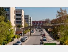 Wohnsiedlung mit mehrstöckigen, modern gestalteten Wohngebäuden, einer von Bäumen und Sträuchern gesäumten Straße und einer Betonbrücke mit der Nummerierung „149-103“ darüber. Entlang der Straße sind parkende Autos und vereinzelte Fußgänger und Radfahrer zu sehen.