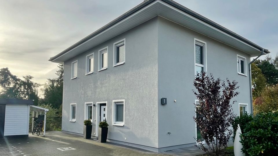 Zweistöckiges, modern wirkendes Wohnhaus mit hellgrauer Fassade, flachem Walmdach und mehreren rechteckigen Fenstern. Vor dem Eingang liegen gepflasterte Stellflächen; links steht ein kleiner Carport, rechts sind Sträucher und Bäume am Grundstücksrand.