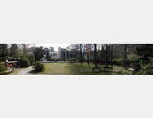 Weitläufiger Garten mit Rasenfläche, Sitzbänken und umgebenden Bäumen, im Hintergrund ein mehrstöckiges Gebäude im historischen Stil mit roten und grauen Fassaden sowie weiteren Wohnhäusern. Eine Ziegelmauer und Laternen säumen den Garten.