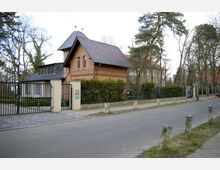 Backsteingebäude mit Satteldach und verzierten Fassadenelementen, umgeben von einer Hecke und einem schmiedeeisernen Zaun. Im Vordergrund führt eine gepflasterte Straße vorbei, gesäumt von Gehweg und schmalen Pollern, während im Hintergrund hohe Bäume stehen.