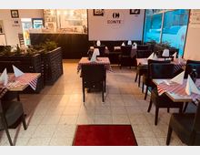 Restaurant-Innenraum mit gefliesten Böden, kleinen Tischen mit rot-weiß karierten Tischdecken und gefalteten Servietten. Im Hintergrund eine Theke mit Regalen voller Flaschen und einer leuchtenden Beleuchtung, sowie große Fenster auf der rechten Seite.