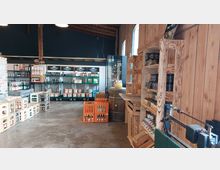 Ein rustikaler Hofladen mit Holzwänden und Regalen, auf denen verschiedene regionale Produkte wie Marmeladen, Getränke und Konserven ausgestellt sind. Im Raum befinden sich Holz- und Plastikboxen, ein Fass sowie eine alte Waage, die den ländlichen Charakter unterstreichen.