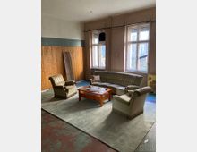 Ein spärlich eingerichteter Raum mit hohen, alten Doppelfenstern, teils abgenutzten Wänden und Holzvertäfelung an einer Seite. In der Mitte steht eine Sitzgruppe aus Sofa und zwei Sesseln auf einem großen Teppich um einen niedrigen Holztisch.