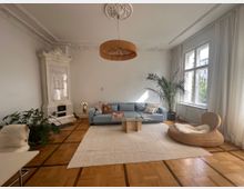 Helles Wohnzimmer mit Holzboden, einer hellblauen Couch, einem weißen Kachelkamin mit verziertem Rahmen, großen Fenstern mit weißen Vorhängen und Zimmerpflanzen. Ein runder Rattansessel mit Fell, ein Glastisch und eine beige Teppichfläche befinden sich ebenfalls im Raum.