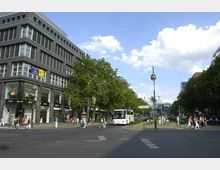 Straßenszene in einer Stadt mit modernen Gebäuden, einem breiten Gehweg, grünen Bäumen und parkenden Bussen am Straßenrand. Im Hintergrund ist eine Freifläche mit einem schlanken Uhrenturm sichtbar, die von Fußgängern und Fahrzeugen genutzt wird.