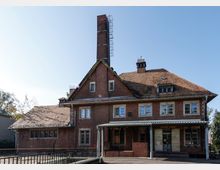 Backstein-Industriegebäude mit steilem Ziegeldach und hoher Schornsteinanlage, vorne ein überdachter Eingangsbereich mit Metallstützen. Mehrere rechteckige Fenster und kleine Dachaufbauten sind sichtbar, davor ein gepflasterter Bereich mit Geländer unter klarem Himmel.