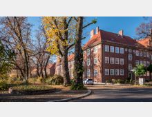 Ein rotes Backsteingebäude mit weißen Fenstern und einem roten Ziegeldach steht an einer baumbestandenen Straße. Im Vordergrund befinden sich ein kleiner bepflanzter Bereich, Parkbänke und herbstlich gefärbte Bäume.
