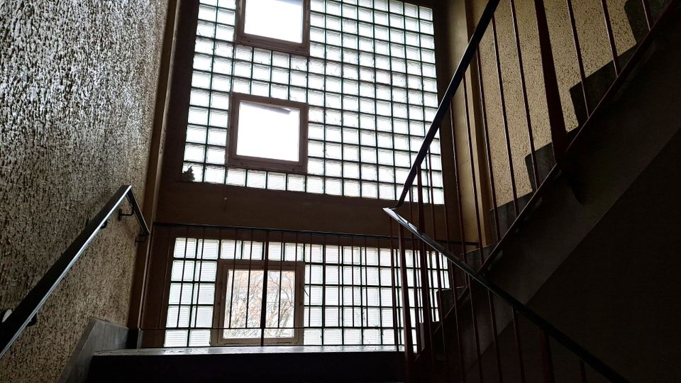 Treppenhaus in einem Mehrfamilienhaus mit rau verputzten Wänden und Metallgeländern, aufgenommen von unten nach oben. Große Glasbausteinfenster und einige rechteckige Klappfenster lassen viel Tageslicht in den Innenraum.