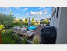 Holzterrasse hinter einem modernen Wohnhaus mit Loungemöbeln, Esstisch, Sonnenschirm und kleinem aufblasbarem Pool, daneben ein abgedeckter Grill. Umgeben von einem bepflanzten Garten mit Hecken und Blick auf benachbarte Häuser unter blauem Himmel mit Wolken.