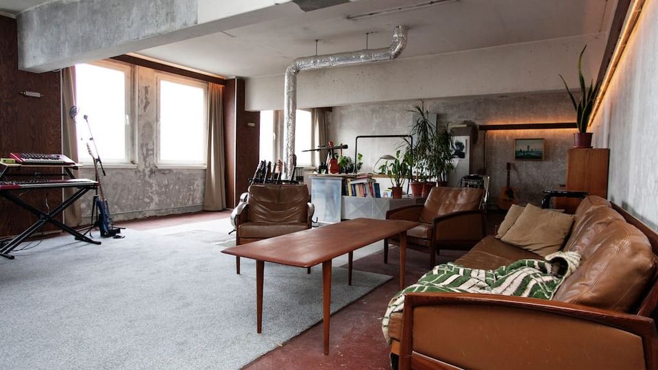 Großer, loftartiger Wohnraum mit rohen Betonwänden, sichtbaren Lüftungsrohren und großen Fenstern mit Vorhängen. Im Vordergrund stehen braune Ledersofas und ein Holztisch auf einem Teppich; im Hintergrund sind Pflanzen, Regale und Musikinstrumente zu sehen.