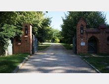 Eingangsbereich mit einer gepflasterten Einfahrt, flankiert von zwei roten Backsteinsäulen mit Spitzbögen und Lattenzauntoren. Im Hintergrund führt ein Weg durch eine grüne Baumallee, die von Sonnenlicht beleuchtet wird.